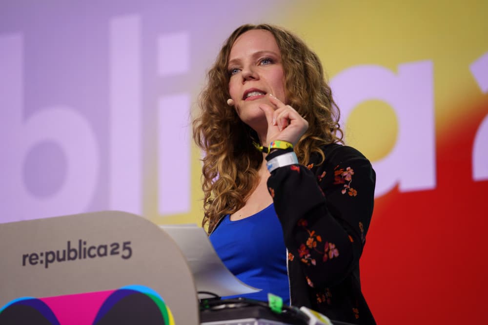 Die Kabarettistin und Autorin Sarah Bosetti steht auf der Bühne der re:publica 2025. Sie trägt ein Mikrofon am Ohr, spricht engagiert ins Publikum und gestikuliert mit der linken Hand. Vor ihr ein Redner:innenpult mit dem Logo der Konferenz. Im Hintergrund ist der bunte „re:publica25“-Schriftzug zu sehen.
