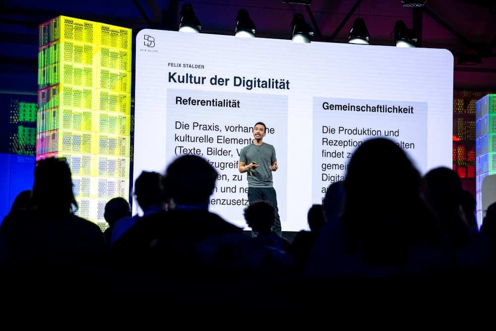 Bob Blume steht auf der Bühne der re:publica 25 vor einer großen Leinwand, die den Titel seiner Präsentation „Kultur der Digitalität“ zeigt. Im Publikum sind die Köpfe der Zuhörenden nur als Silhouetten zu erkennen, während Blume in T-Shirt und Jeans, ein Mikrofon in der Hand hält. 