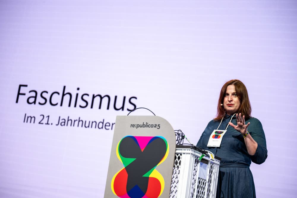 Die Politikwissenschaftlerin Natascha Strobl steht auf einer Bühne der re:publica 25. Sie trägt ein dunkles Kleid, hat schulterlanges rötlich-braunes Haar und spricht in ein Headset-Mikrofon. In der linken Bildhälfte ist die große Projektion ihres Vortragstitels zu sehen: „Faschismus im 21. Jahrhundert“. Vor ihr steht ein Rednerpult mit dem re:publica-Logo. Strobl hebt die rechte Hand gestikulierend in einer erklärenden Bewegung. Die Stimmung wirkt ruhig und konzentriert.