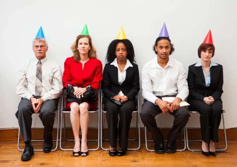 Fünf Personen mit Büro Outfits und Partyhütchen sitzen nebeneinander aufgereiht auf Stühlen, Gesichter neutral