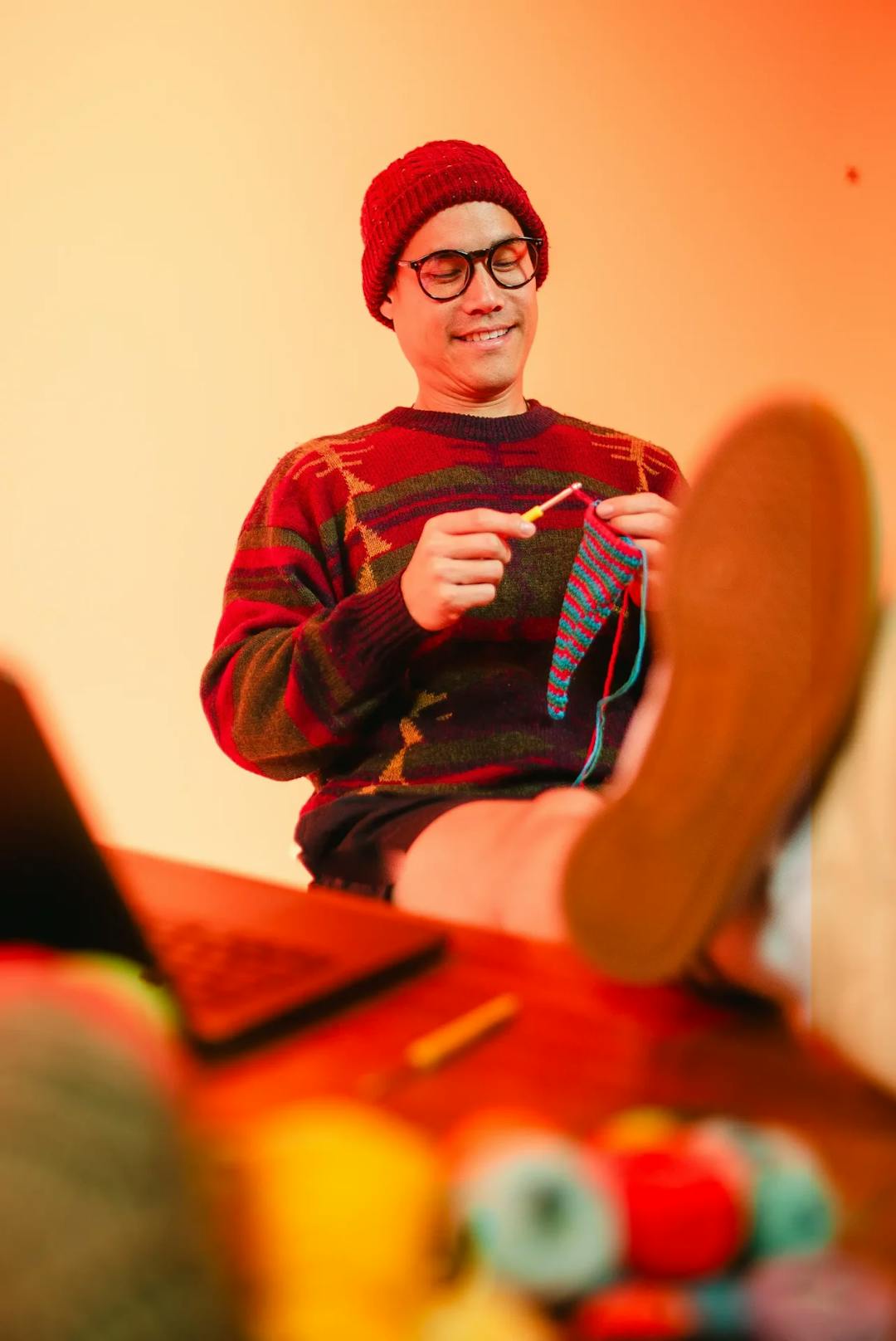 Ein lächelnder Mann mit gelb-rotem Strickpullover, Brille und Mütze sitzt an einem Schreibtisch, hat die Füße neben seinem Laptop auf der Tischplatte und strickt.