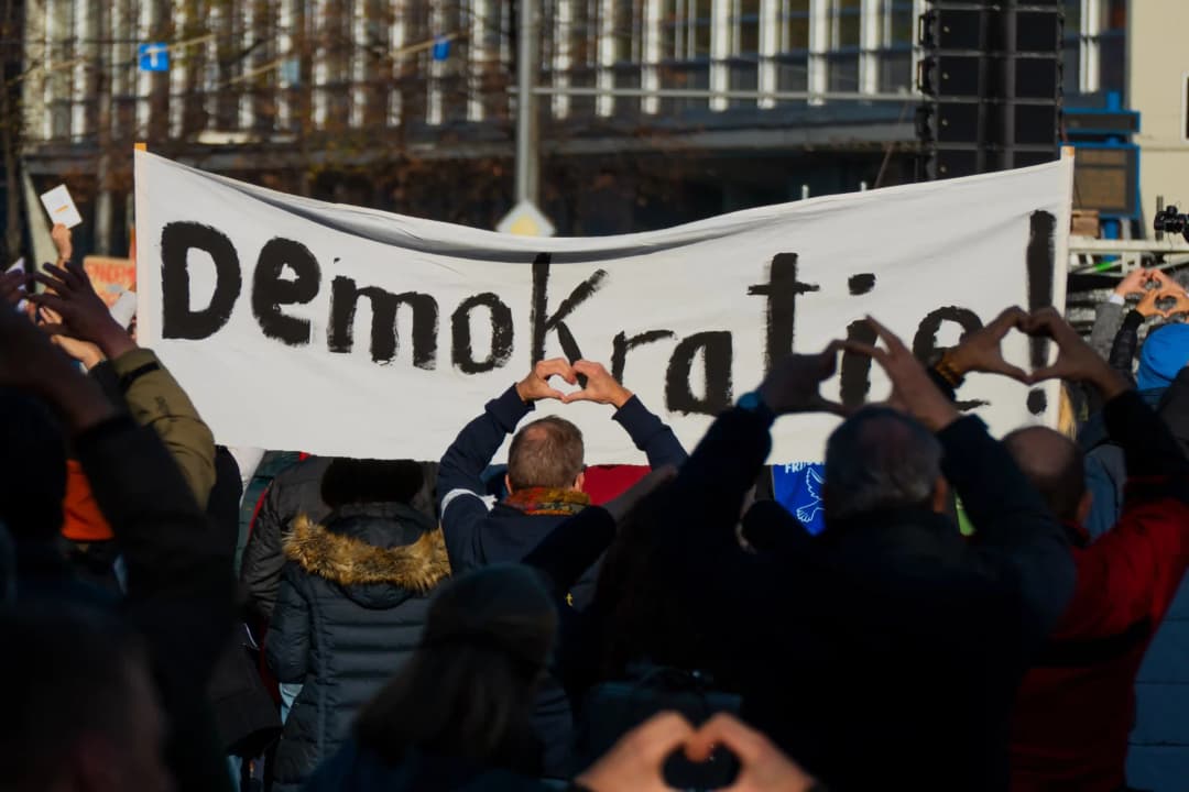 Menschen demonstrieren für die Demokratie. Sie halten eine Fahne, auf der Demokratie zu lesen ist und formen die Hände zu Herzen, die sie in die Höhe halten.