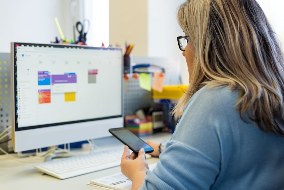 Frau sitzt am Schreibtisch vor einem Bildschirm und schaut auf ihr Smartphone. 