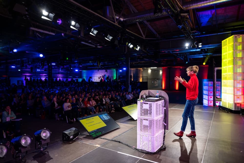 Maja Göpel auf der re:publica 2025: „Digitalisierung ist ein Gestaltungsprojekt, kein Naturereignis“ Maja Göpel steht auf der Bühne der re:publica 25 und hält einen Vortrag. Sie trägt einen roten Rollkragenpullover, eine blaue Jeans und rote Stiefeletten. Ihre Haare sind zu einem lockeren Dutt zusammengebunden. Sie spricht gestikulierend ins Publikum, das in dunklem Licht vor ihr sitzt und gespannt zuhört. Vor ihr auf der Bühne stehen ein Rednerpult aus weißen Kunststoffkisten sowie zwei große Monitore mit Stichpunkten zur Präsentation. Die Halle ist mit farbigen Lichtern in Blau, Lila und Gelb atmosphärisch ausgeleuchtet.