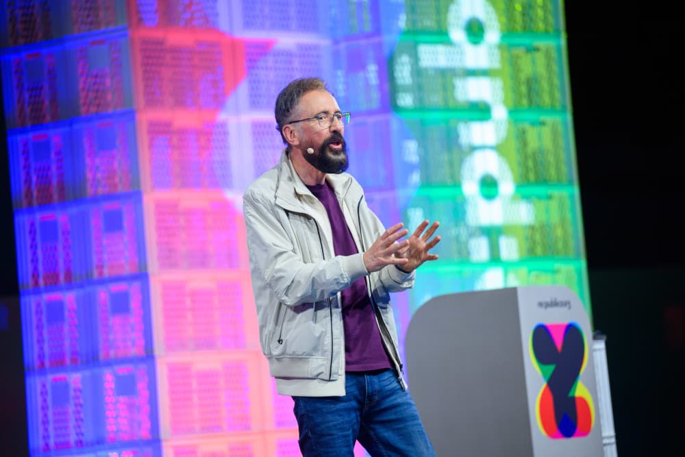 André Frank Zimpel auf der re:publica 25: Mehr Sichtbarkeit für neurodivergente Perspektiven André Frank Zimpel steht auf der Bühne der re:publica 25 und spricht mit lebhafter Gestik ins Publikum. Er trägt eine helle Jacke über einem violetten T-Shirt und eine blaue Jeans. Seine Hände sind erhoben, seine Finger gespreizt, als wolle er einen Gedanken unterstreichen. Er trägt Brille und hat einen dunklen Vollbart. Hinter ihm leuchten bunte Lichtinstallationen aus gestapelten Kunststoffkisten in Pink, Blau, Grün und Gelb. Rechts neben ihm steht ein Rednerpult mit dem Logo der re:publica.