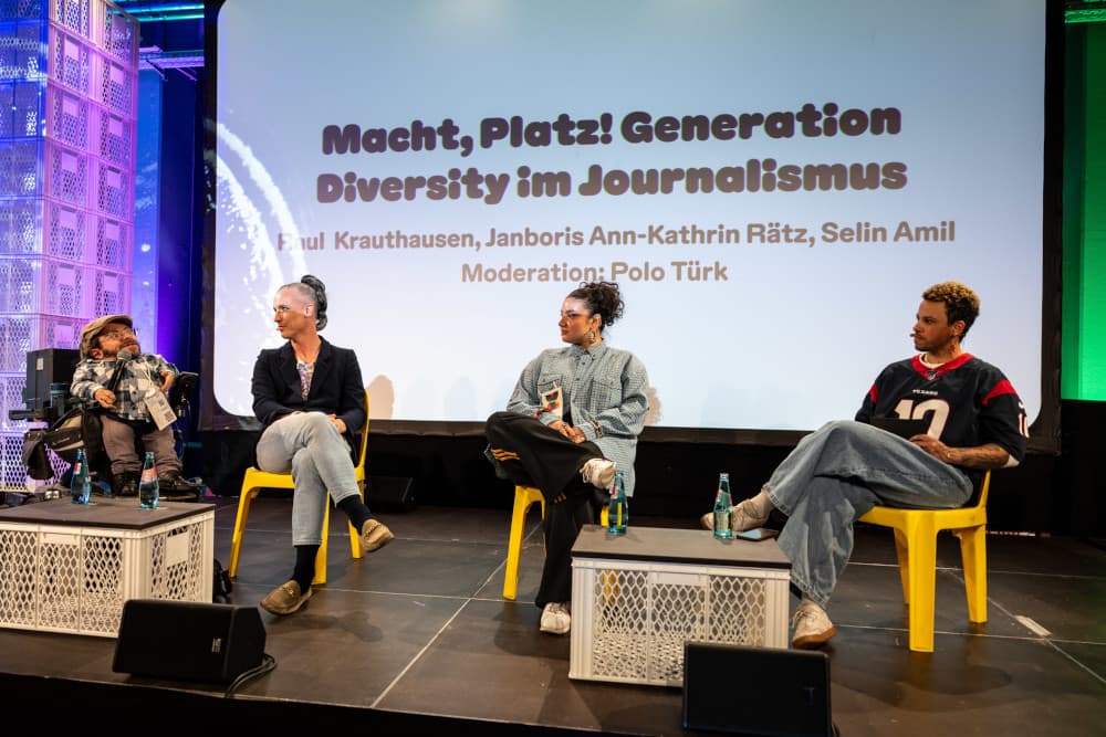 „Macht, Platz!“ – Gerechtere Strukturen im Journalismus im Fokus der re:publica 25 Vier Personen sitzen auf der Bühne der re:publica 25 vor einer Projektion mit dem Titel: „Macht, Platz! Generation Diversity im Journalismus“. Von links nach rechts sind zu sehen: Raul Krauthausen im Rollstuhl mit Mikrofon in der Hand, Janboris Ann-Kathrin Rätz mit geflochtenem Haar und Blazer, Selin Amil mit lockigem Dutt und weitem Hemd sowie Moderator Polo Türk im Football-Trikot. Die Bühne ist farbenfroh ausgeleuchtet, auf dem Boden stehen Wassergläser, ein Mikrofonkabel und improvisierte Tische aus Kunststoffboxen.