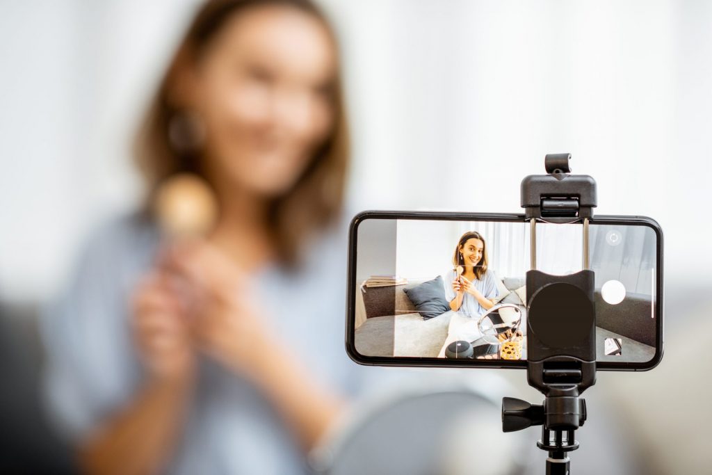 Eine Handykamera filmt eine junge Frau, die ein Produkt in die Kamera hält.  